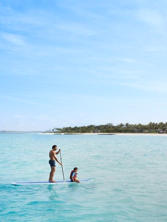Two people on a single paddleboard in clear blue water near a white sand beach during luxury Family vacations.