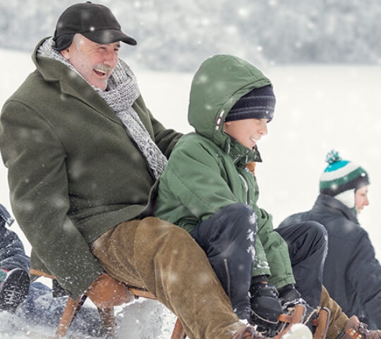 An older man and children laugh while riding sleds down a snowy hill on their luxury Family holidays.