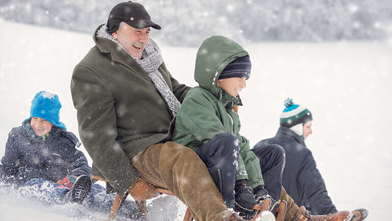 An older man and children laugh while riding sleds down a snowy hill on their luxury Family holidays.