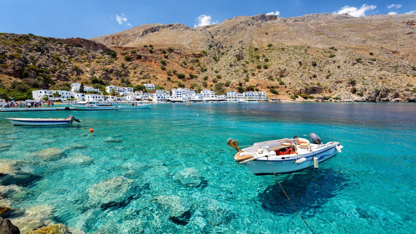 Little boats floating on crystal clear water in a bay next to a small Greek village with white sugar-cube buildings