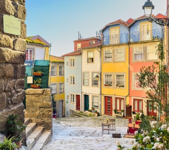 A narrow cobblestone street featuring multi-story buildings painted in various bright colors with small balconies and plants.