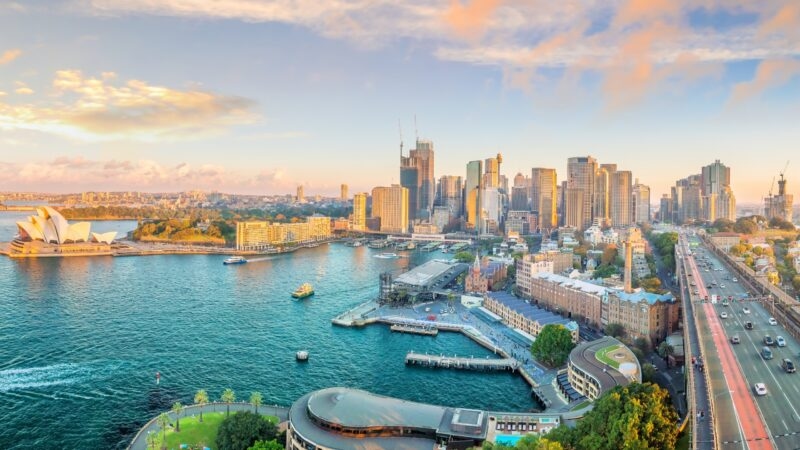 High-angle view of Sydney Harbour featuring the Opera House, city skyscrapers, and a bridge under a sunset sky.