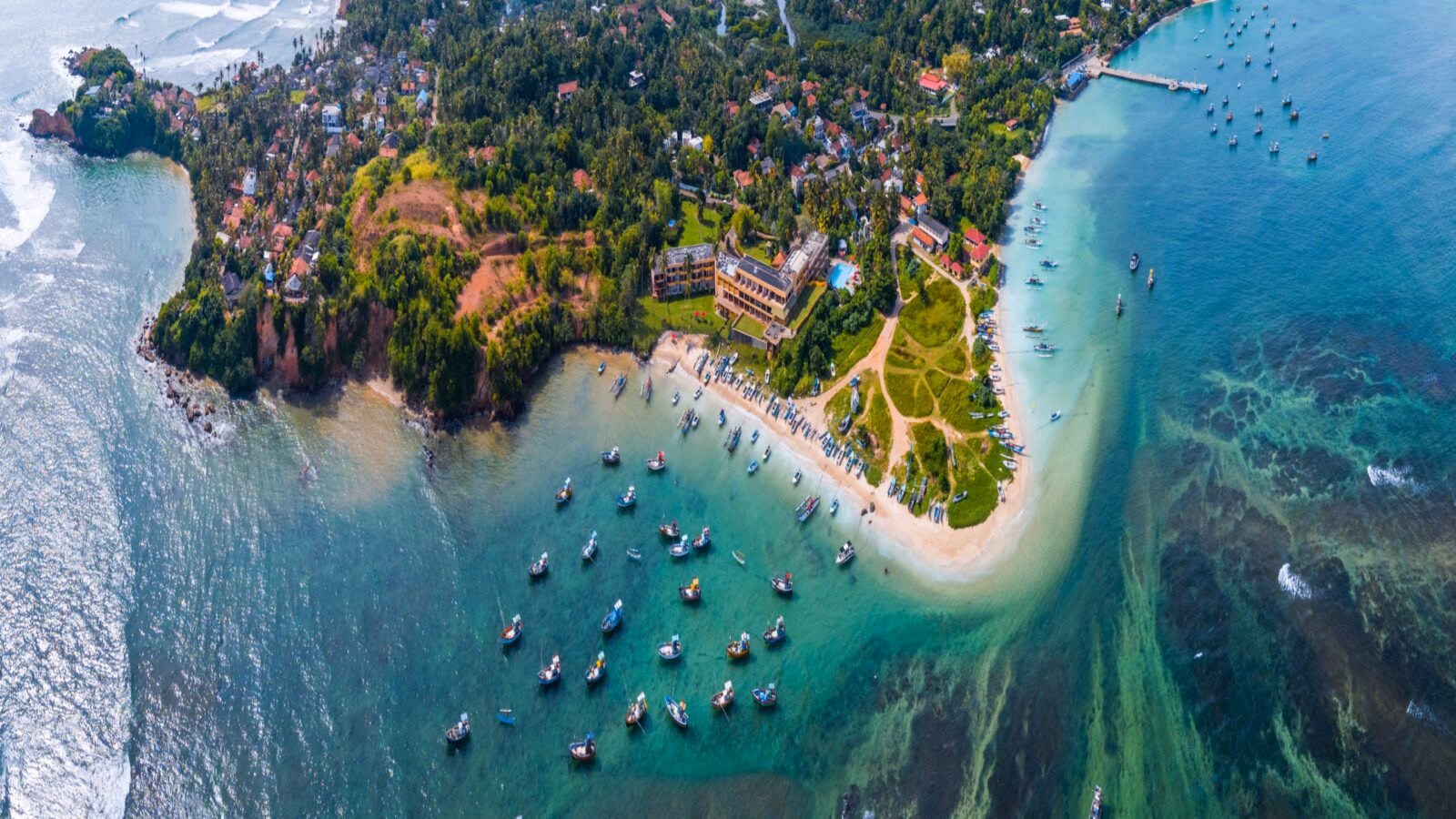 Aerial view of a tropical coast with a lush village, sandy beach, and many small boats in turquoise water.