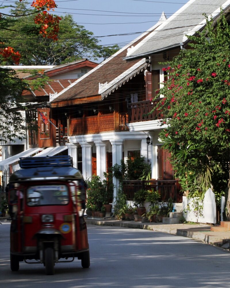 A red tuk-tuk travels down a quiet street lined with traditional houses during luxury Laos holidays.