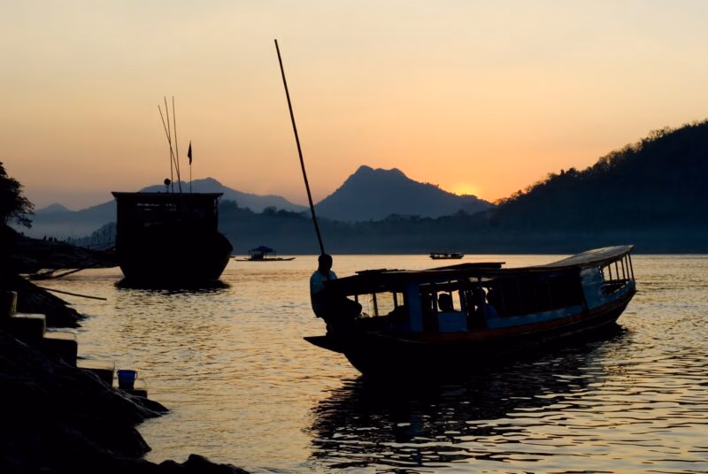 Silhouetted boats on the Mekong River at sunset with mountain backdrops during luxury Laos holidays.