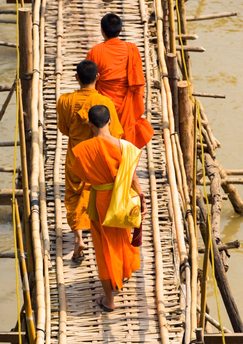 Three monks wearing orange robes cross a rustic bamboo bridge, a serene scene from luxury Laos holidays.