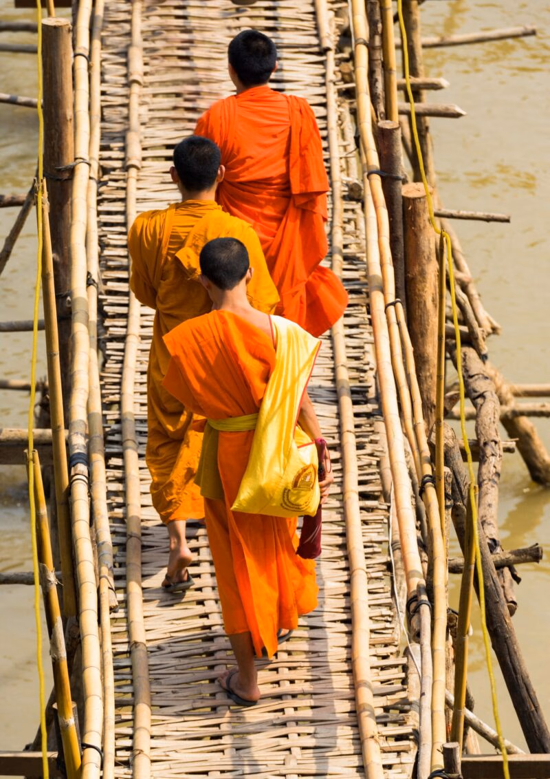 Three monks wearing orange robes cross a rustic bamboo bridge, a serene scene from luxury Laos holidays.