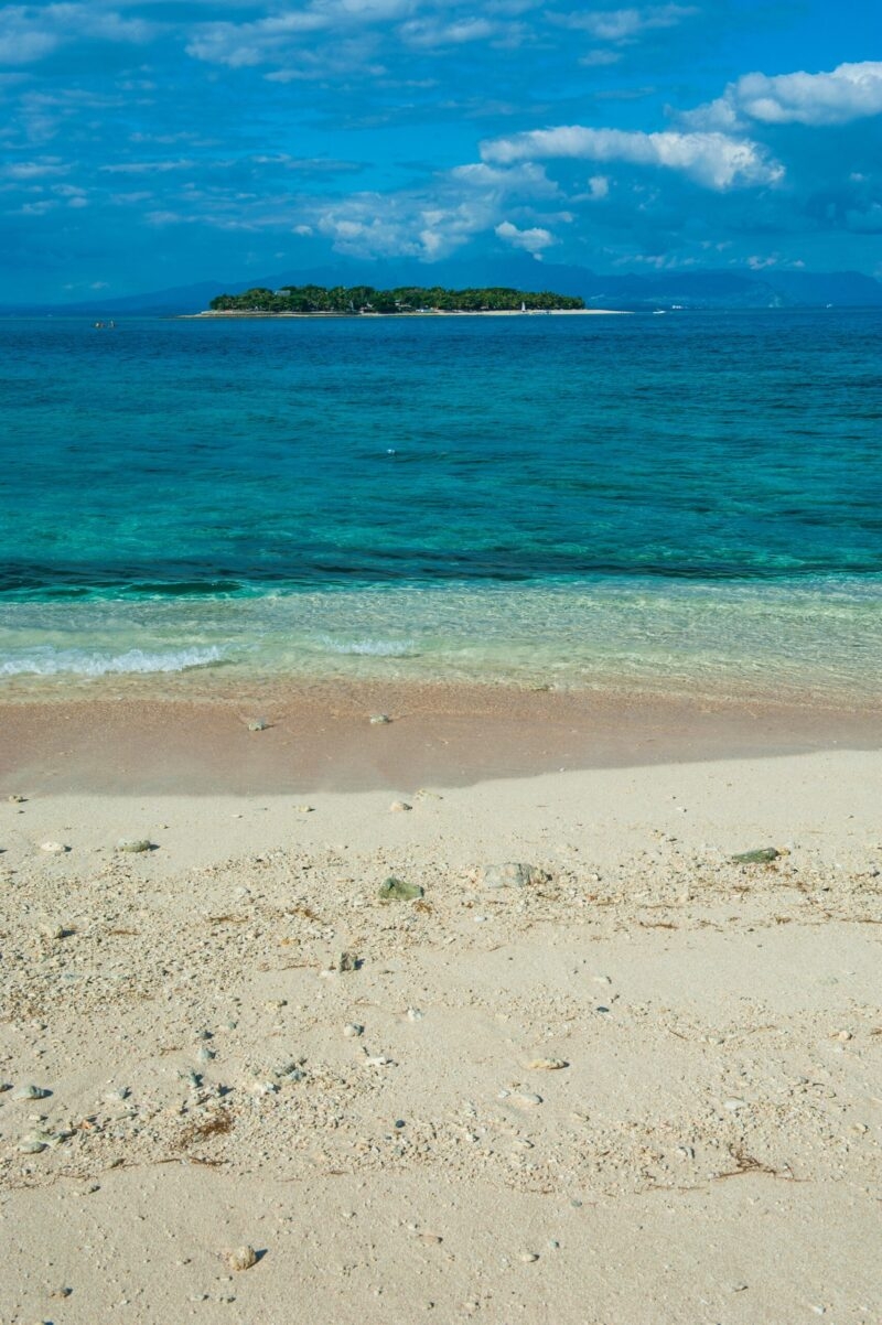 Sandy shoreline looking out over the blue sea toward a small forested island under clouds during luxury Fiji vacations.
