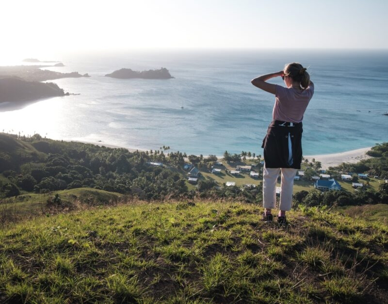 Woman standing on a grassy hill looking out over a tropical bay and village for luxury Fiji tours.