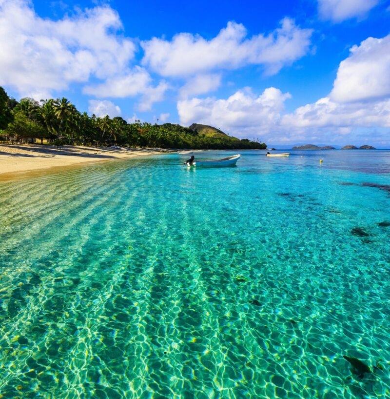 Clear shallow turquoise water on a sandy beach with palm trees and a small boat for luxury Fiji tours.