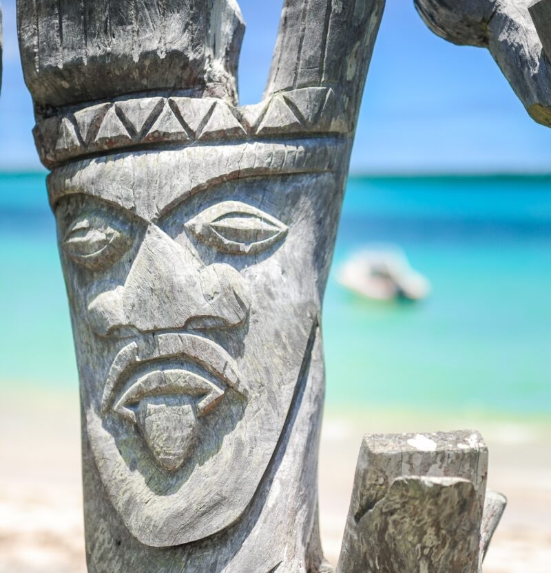 Close-up of a traditional wooden face carving on a beach with the tropical ocean behind for luxury Fiji vacations.