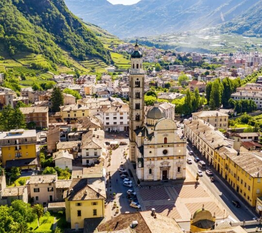 High-angle view of a town with a prominent stone church tower and surrounding buildings in a green valley.