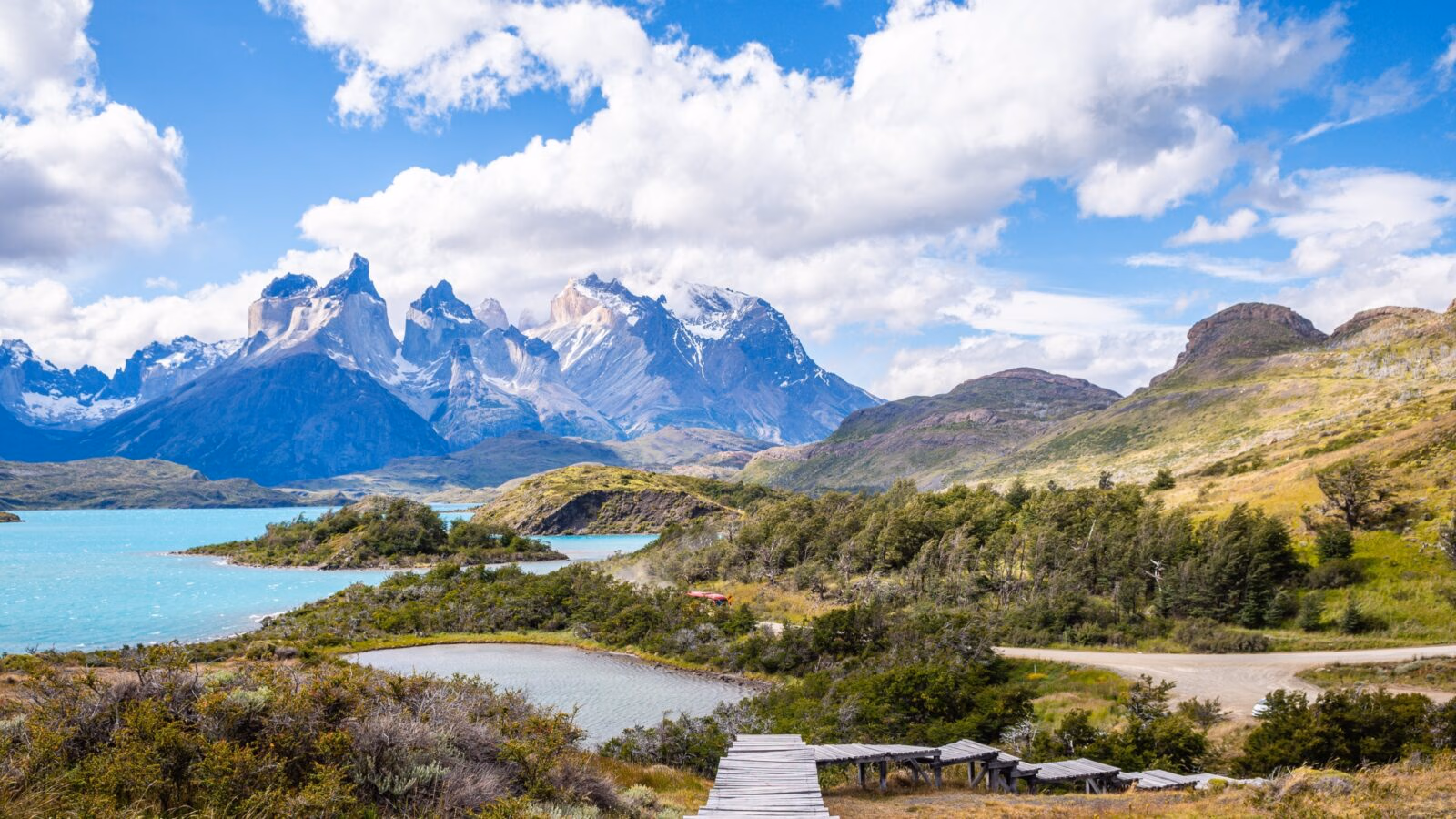Dramatic mountain peaks rise above a bright blue lake and green hills under a cloudy sky in Torres del Paine.