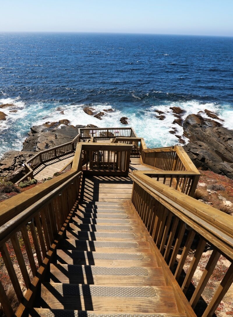 View from the top of wooden stairs leading down to a rocky coastline with waves hitting the shore.