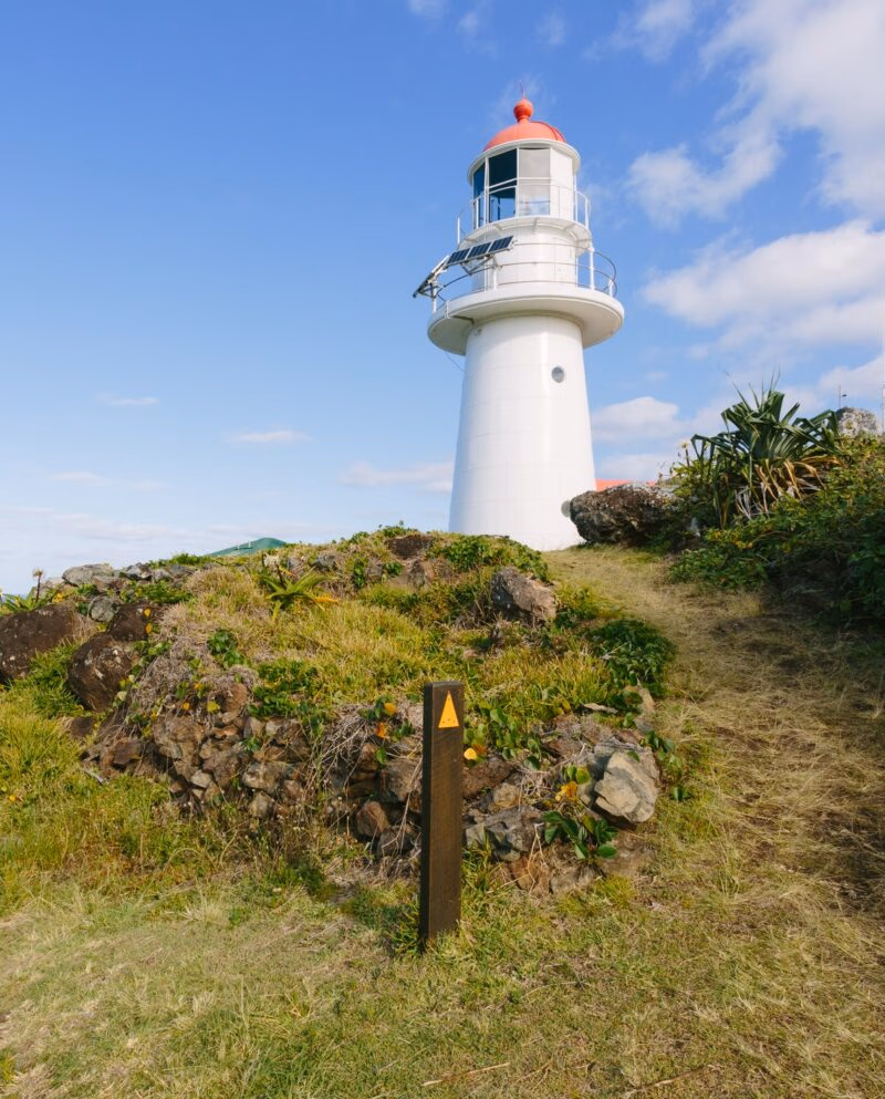 A white lighthouse with a red top situated on a grassy hill with a small path and a wooden trail marker.