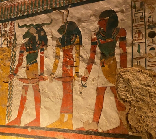Ancient Egyptian wall art showing three figures with animal heads and rows of hieroglyphics on a textured surface.