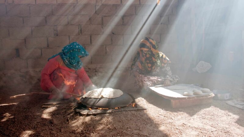Two people cooking flatbread on a large domed griddle over a fire in a dusty, sunlit courtyard.