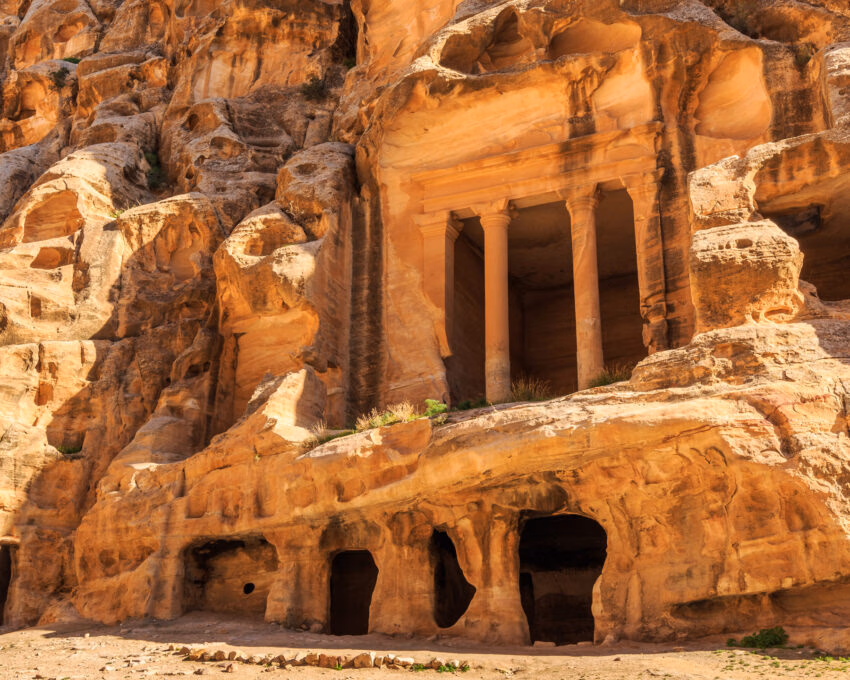 Caved buildings of Little Petra in Siq al-Barid, Wadi Musa, Jordan. Large ancient rock-cut tomb with tall columns and dark cave openings carved into a textured tan sandstone cliff.