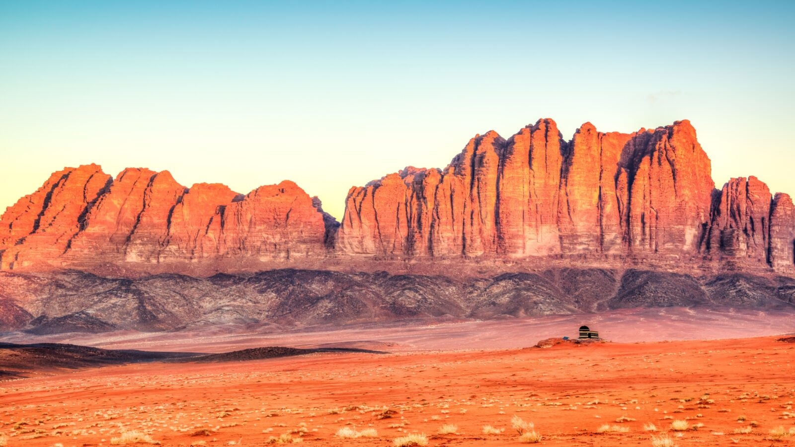 A wide shot of dramatic red rock mountains rising from a flat desert floor at sunset.