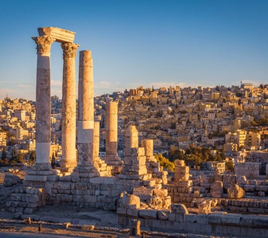 Ancient Roman columns of the Temple of Hercules overlook a hillside city in Amman, Jordan, during sunset.