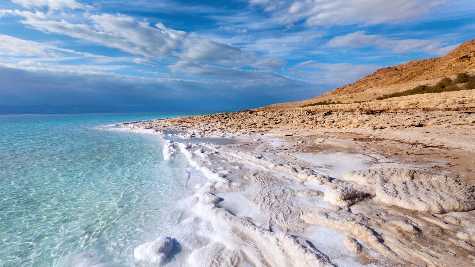 The Dead Sea shoreline featuring bright white salt formations, clear turquoise water, and rocky, beige hills under a blue sky.