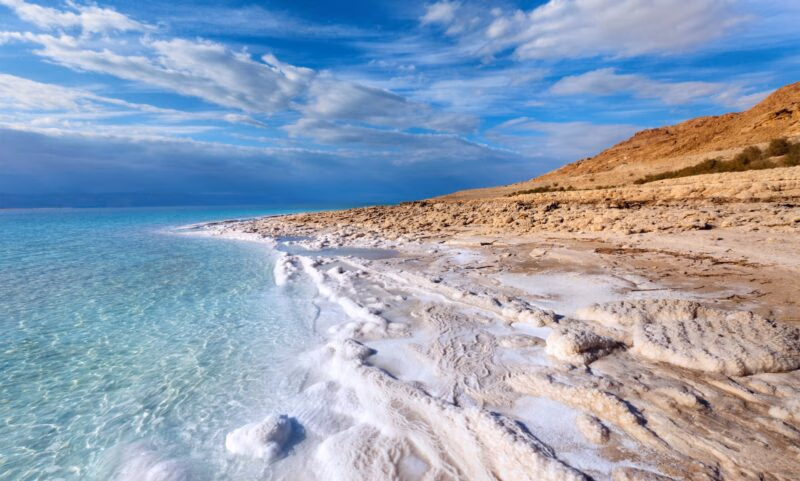 The Dead Sea shoreline featuring bright white salt formations, clear turquoise water, and rocky, beige hills under a blue sky.