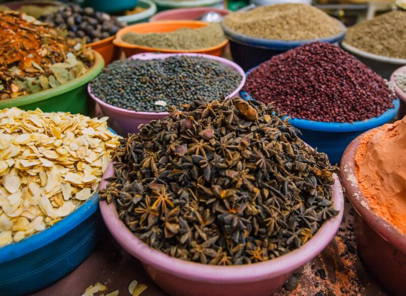 Close-up of various spices and grains, including star anise, lentils, dried garlic flakes, and red seeds, displayed in market bowls.