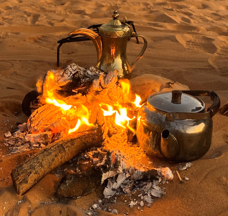 Two brass kettles, a traditional coffee pot and a round tea pot, sit beside a small campfire on the desert sand.
