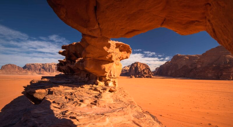 Natural sandstone rock arch formation frames the reddish-orange desert and mountains of Wadi Rum under a deep blue sky.
