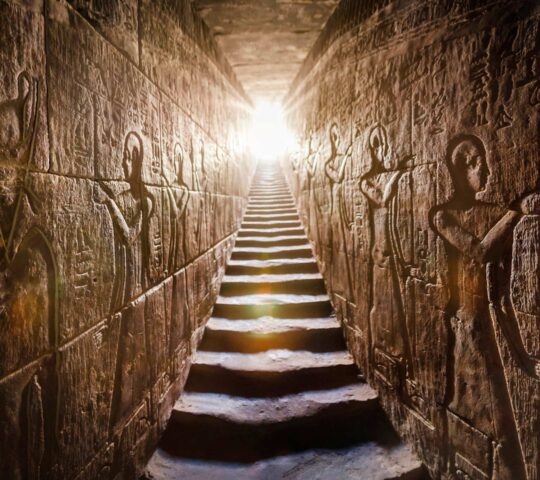 A stone stairway between two walls featuring intricate Egyptian reliefs leading up toward a bright light.