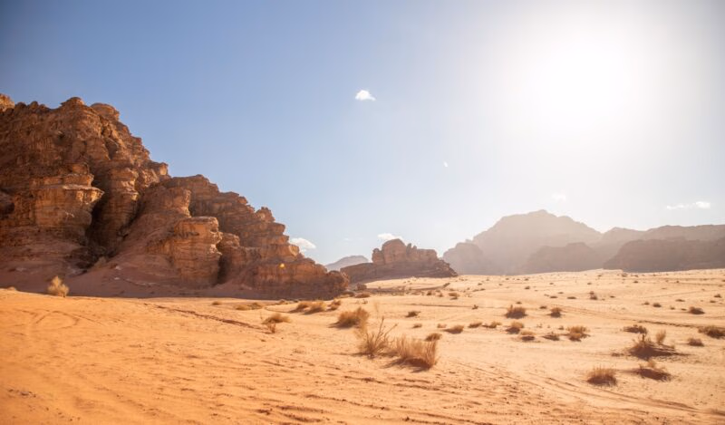 A wide, bright view of a red-sand desert with tall, rugged sandstone mountains under a clear blue sky. Luxury Middle East trips.