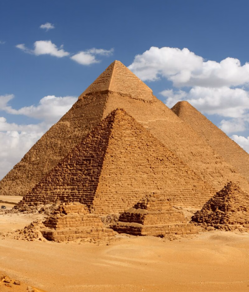 Three immense, ancient pyramids rise from the sandy desert ground under a blue, cloudy sky. An essential sight for a luxury Middle East vacation.