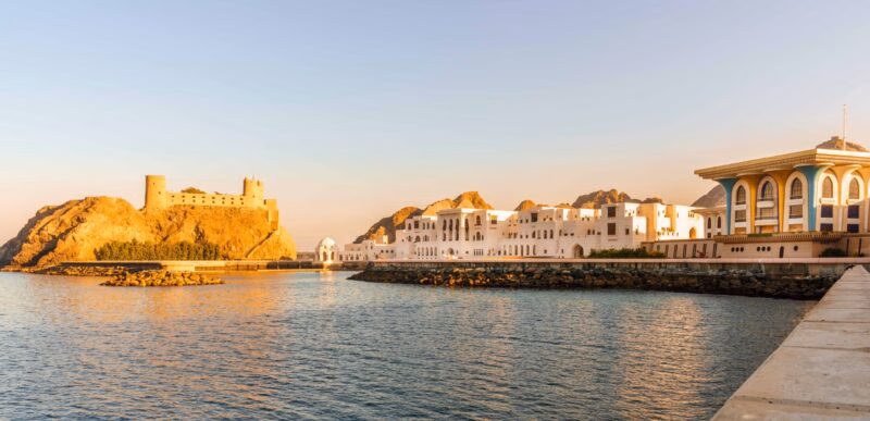 A scenic coastal view of a fort and white palace buildings at sunset on luxury Oman tours.