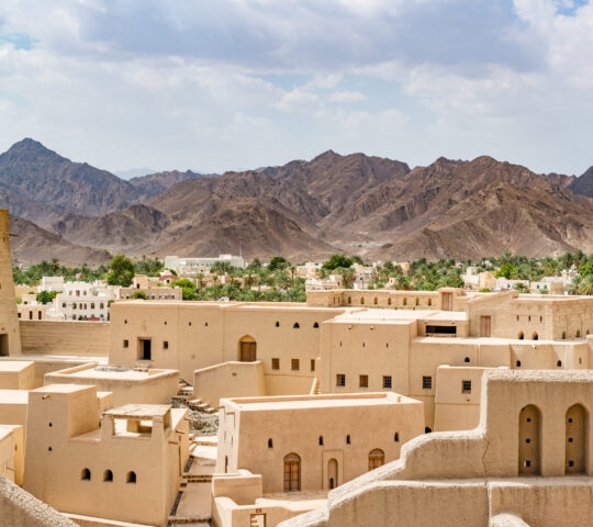 An ancient tan stone fort complex with a round tower and mountain backdrop during luxury Oman vacations.