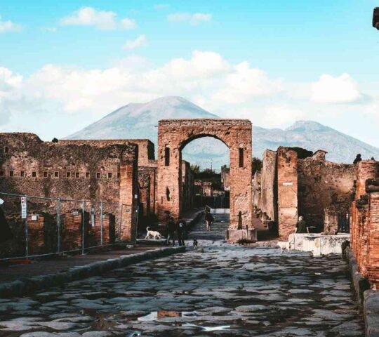 A view down a stone path lined with ruined brick walls leading toward a large mountain under a cloudy sky.