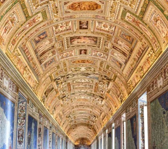 A long hallway with a highly decorated arched gold ceiling and large blue maps painted on the side walls.