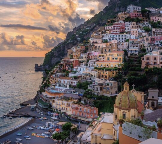 Sunset over a colorful coastal town built into a steep mountain cliff overlooking the sea.