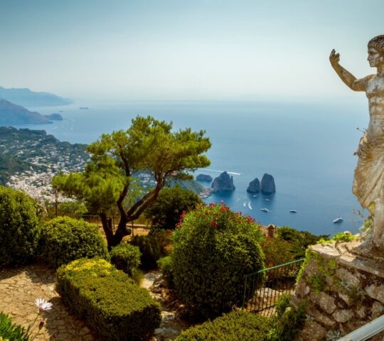 A stone statue stands on a terrace overlooking the blue sea and distant rocky islands in Capri.
