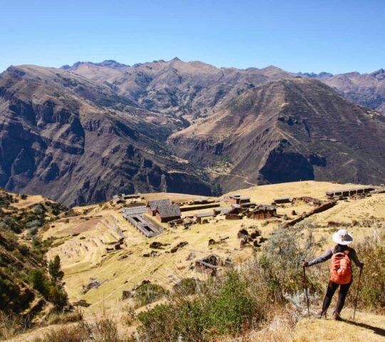 Hiker with an orange backpack and white hat overlooking the Inca ruins of Pisac on a steep, arid mountain slope in Peru.