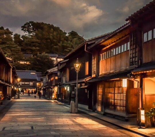 A charming evening scene of a traditional Japanese street. The cobblestone pathway is lined with historic wooden buildings, their warm, glowing lanterns illuminating the surroundings. The sky is dusky, with hints of clouds adding depth to the serene atmosphere. Soft light from the windows and lanterns casts a cozy and inviting glow, reflecting the timeless beauty and cultural heritage of the area. In the background, trees and additional buildings are visible, completing the picturesque setting.