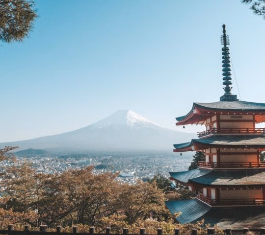 A five-story red pagoda overlooks a valley with the snow-capped peak of Mount Fuji under a clear blue sky.