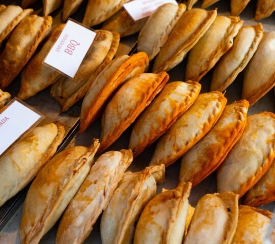 Rows of golden-brown baked empanadas are arranged in a display case with labels like BBQ.