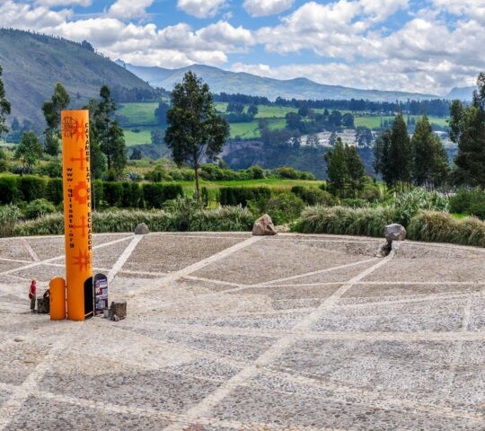 An orange pillar monument stands in the center of a paved circular area with green mountains in the distance.