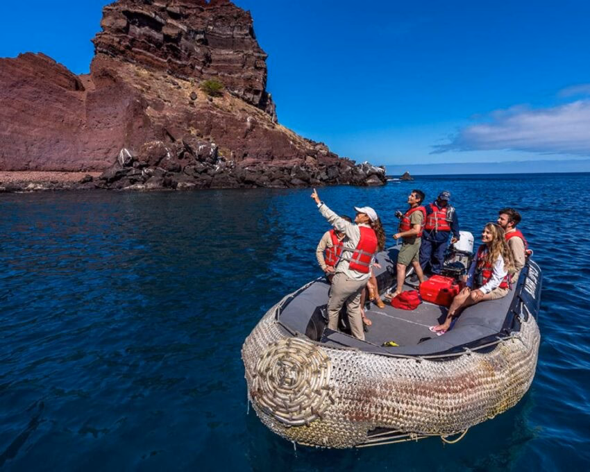People wearing life jackets ride in a small boat near a tall, reddish-brown rock cliff in the ocean.