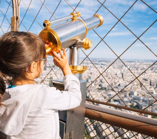 A young girl looks through a gold-colored telescope at the city of Paris from an observation deck.