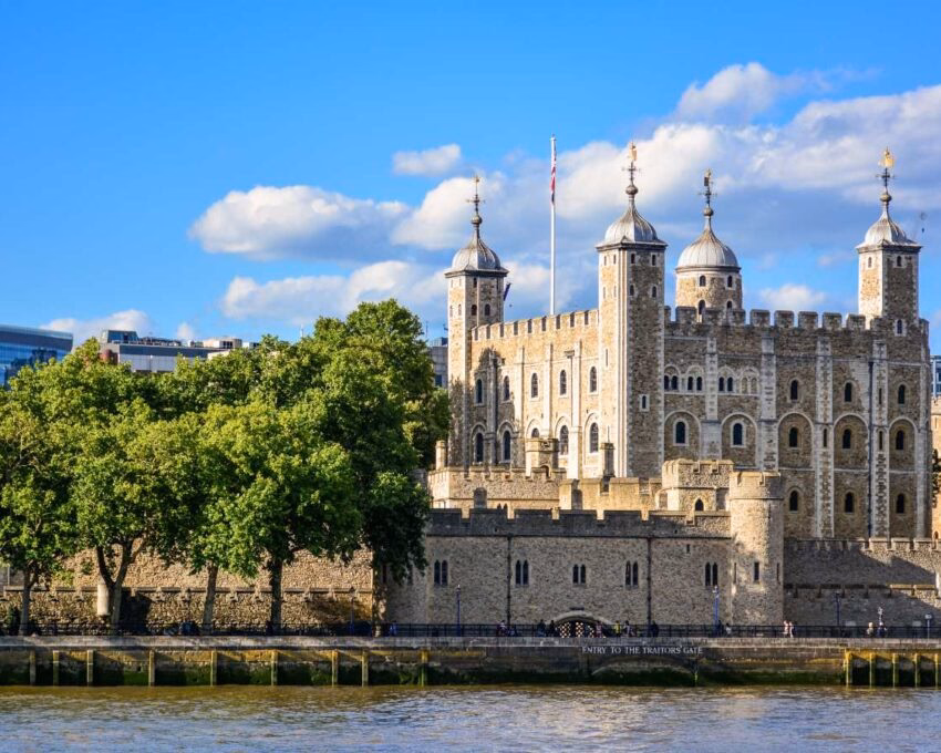 The stone towers of the Tower of London viewed from across the River Thames behind a line of green trees.