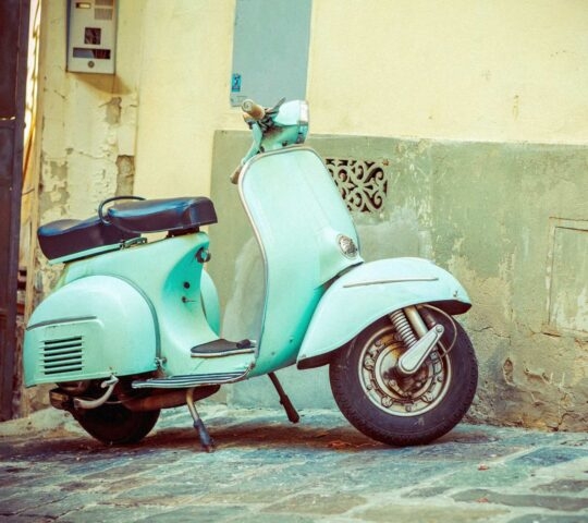 A light blue-green vintage scooter leaning on its kickstand on a stone-paved street next to a building.