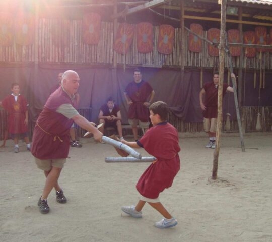 An adult and child in Roman tunics practice sword fighting in a training arena.