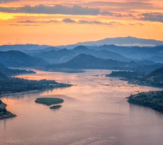 Wide river winding through mist-covered mountains under a vibrant orange and yellow sunset sky.