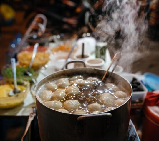 Close-up of a large pot of boiling broth and meatballs with visible steam rising, set against a blurred background.